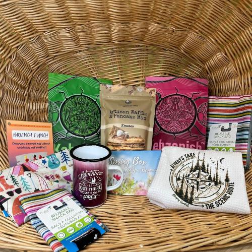 The contents of the Spring 2024 Glamping Box: Brunch Punch recipe card, Kebaonish Tea in two varieties, Colibri reusable snack bags in three sizes, a white tea-towel with the wording "always take the scenic route" and a burgundy "adventure is out there - let's go find it" enamel mug.