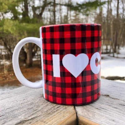 This is a picture of our 12 oz Glamping Ceramic Coffee Mug.  The mug has a Buffalo Check background (red and black plaid pattern) with 'I Heart Coffee' written in a large white font.  The ceramic mug sits on a wooden railing overlooking the woods, living its best coffee mug life.