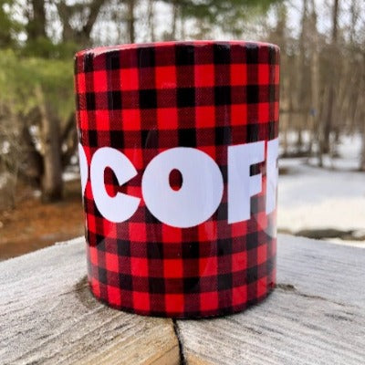 This is a picture of our 12 oz Glamping Ceramic Coffee Mug.  The mug has a Buffalo Check background (red and black plaid pattern) with 'I Heart Coffee' written in a large white font.  The ceramic mug sits on a wooden railing overlooking the woods, living its best coffee mug life.