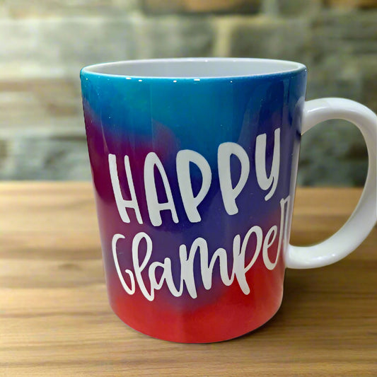 Ceramic mug with a blue, purple and red tie-dye background, the words "Happy Glamper" are written in white font.  The mug sits upon a workbench.