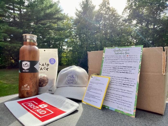 A picture of the September Glamping Box in its entirety: maple praline almonds, water bottle, first aid kit pouch, 'Worry Less Wander More' ball cap in grey. Plus a drink recipe and info card.