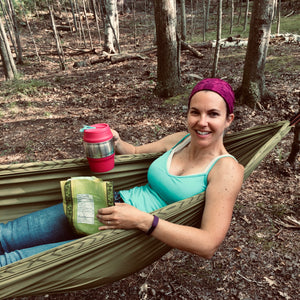 This is a picture of our founder in her natural element... Glamping in a green hammock holding a pink insulated drink holder and a snack, smiling with forest in the background.  She is living her best live.  Glamp on!
