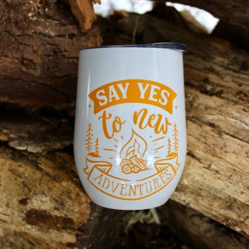 This is a picture of our Glampy Wine Tumbler.  The wine tumbler is white with the saying "Say YES to Adventure" printed on it in the colour 'goldenrod' like a dark yellow or light orange.  The tumbler is nested amongst corded wood, livin' it's best glamping life.