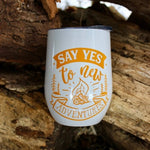 Load image into Gallery viewer, This is a picture of our Glampy Wine Tumbler.  The wine tumbler is white with the saying &quot;Say YES to Adventure&quot; printed on it in the colour &#39;goldenrod&#39; like a dark yellow or light orange.  The tumbler is nested amongst corded wood, livin&#39; it&#39;s best glamping life.

