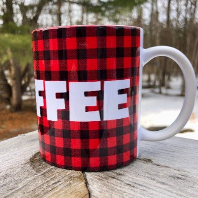 This is a picture of our 12 oz Glamping Ceramic Coffee Mug.  The mug has a Buffalo Check background (red and black plaid pattern) with 'I Heart Coffee' written in a large white font.  The ceramic mug sits on a wooden railing overlooking the woods, living its best coffee mug life.
