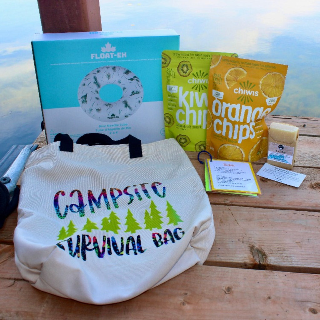 The complete July Glamping Box containing a Float-Eh water tube, two bags of Chiwis natural fruit snacks (kiwi and orange), a campsite survival canvas bag, LaMer natural soap, drink recipe, info card and more!  All pictured on a dock in the summer.