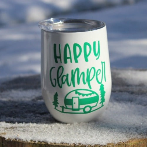 This is a picture of our Glampy Wine Tumbler. The wine tumbler is white with the saying "Happy Glamper" printed on it in green. The tumbler is peacefully sitting on a snow covered wooden stump, livin' it's best glamping life.