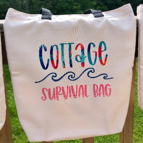 Glamping Essentials' 'Cottage Survival Bag' in neutral beige with Cottage in a tropical flower font and Survival Bag in pink striped font below a row of blue waves.  Bag had two black handles and is draped over a rail in the woods.