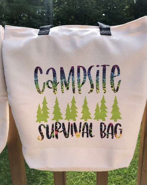 Glamping Essentials' 'Campsite Survival Bag' in neutral beige with Campsite in a rainbow font and Survival Bag in black font with flowers below a row of green pine trees.  Bag had two black handles and is draped over a rail in the woods.