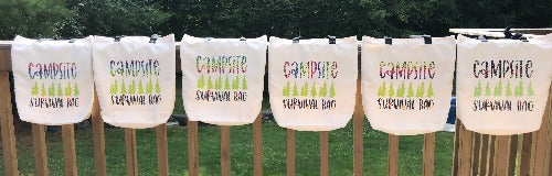 A row of Glamping Essentials' 'Campsite Survival Bag' in neutral beige with Campsite in a rainbow font and Survival Bag in black font with flowers below a row of green pine trees.  Bag had two black handles and is draped over a rail in the woods.