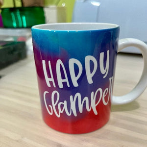 This is a ceramic mug with a blue, purple and red tie-dye background, the words "Happy Glamper" are written in white font.  The mug sits upon a workbench.