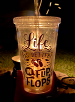 Load image into Gallery viewer, From the June Glamping Box - a picture of the clear plastic tumbler with a straw that says &quot;Life is Better in Flip Flops&quot; in shimmery purple font. The cup is pictured here at night in front of the campfire.
