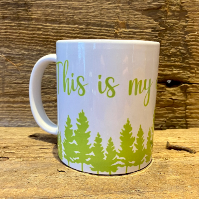 This is our "This is my Cottage / Camping Mug".  The image shows a white ceramic mug with the words "This is My" printed in green above a green pine forest.  The mug is backed against wooden planks.