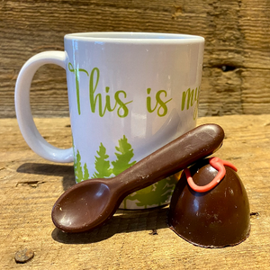 The Glamping Essentials ceramic coffee mug in white with "This is my camping/cottage mug" in green font above a forest of green trees.  In front of the mug is a dark hot chocolate bomb with a dark chocolate mixing spoon.