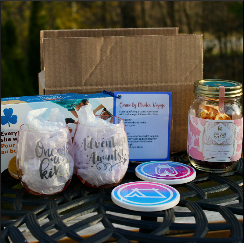 Our May Box 2022 shows one box of Girl Guide cookies, two light weight pink stemless wine tumblers, one that says "One of a Kind" and the other says "Adventure Awaits", plus two coasters, a cocktail infusion jar and a recipe card for a Cosmo cocktail, all nestled in front of a cardboard box.  Cheers!
