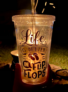 From the June Glamping Box - a picture of the clear plastic tumbler with a straw that says "Life is Better in Flip Flops" in shimmery purple font. The cup is pictured here at night in front of the campfire.