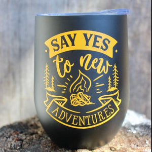 This is a picture of our Glampy Wine Tumbler. The wine tumbler is black with the saying "Say YES to Adventure" printed on it in the colour 'goldenrod' like a dark yellow or light orange. The tumbler is resting on a rock, outdoors, livin' it's best glamping life.