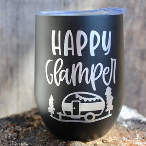 This is a picture of our Glampy Wine Tumbler. The wine tumbler is black with the saying "Happy Glamper" printed on it in silver. The tumbler is resting on a rock, outdoors in the sunshine, livin' it's best glamping life.