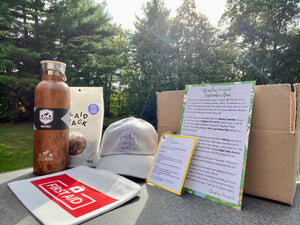 A picture of the September Glamping Box in its entirety: maple praline almonds, water bottle, first aid kit pouch, 'Worry Less Wander More' ball cap in grey.  Plus a drink recipe and info card.